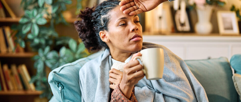 Person drinking tea with other person checking forehead temperature with hand