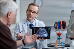 Middle aged Caucasian male urologist explaining ultrasound results to senior Caucasian man using digital tablet, anatomical kidney model and computer monitor visible in medical office