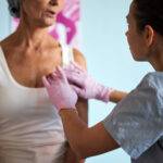 Middle aged Caucasian woman sitting while young adult Caucasian female healthcare worker wearing gloves palpating chest during medical examination in clinical setting related to cancer