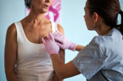 Middle aged Caucasian woman sitting while young adult Caucasian female healthcare worker wearing gloves palpating chest during medical examination in clinical setting related to cancer