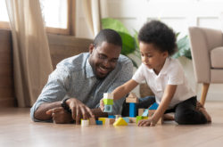 Mixed race young father and small adorable sweet son having fun at modern apartment living room play holding colorful toy blocks set lying sitting on wooden warm floor, family activity at home concept