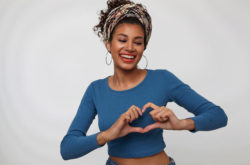 Glad young lovely dark haired curly woman with casual hairstyle smiling happily while forming heart with raised hand, standing against white background