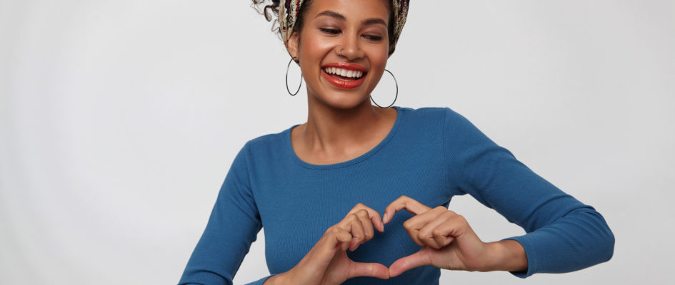 Glad young lovely dark haired curly woman with casual hairstyle smiling happily while forming heart with raised hand, standing against white background