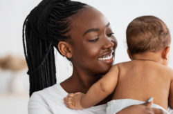 Happy Maternity Concept. Portrait of cheerful African American woman with long afro braids holding and hugging her small kid in diaper, enjoying pastime with lovely baby indoors, blurred backround