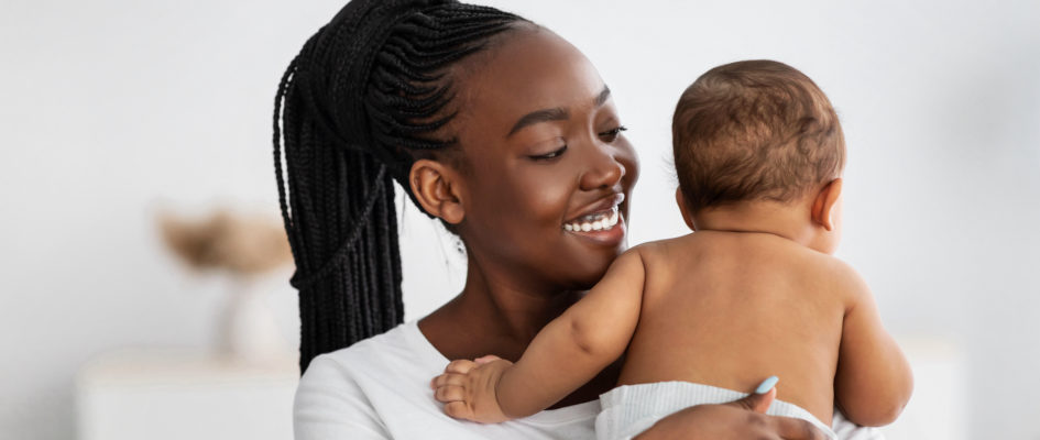 Happy Maternity Concept. Portrait of cheerful African American woman with long afro braids holding and hugging her small kid in diaper, enjoying pastime with lovely baby indoors, blurred backround
