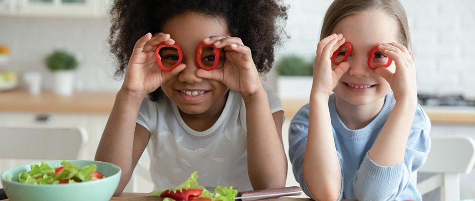 Close up head shot smiling diverse little girls holding red pepper as glasses, sitting at table with vegetables in kitchen, looking at camera, kids multiracial sisters posing for funny portrait